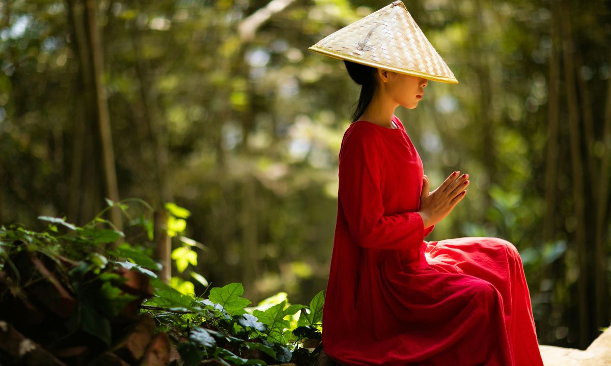 donna asiatica con le mani giunte e un cappello vietnamita seduta ai piedi di un albero