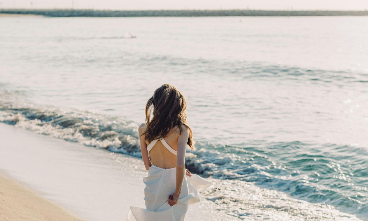 Donna con abito bianco che guarda il mare da una spiaggia luminosa